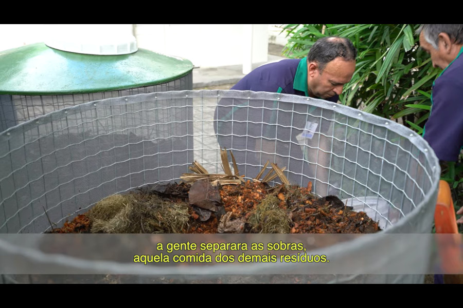 Foto mostra tela do vídeo sobre compostagem, com dois homens colocando resíduos orgânicos em um grande recipiente já com outros resíduos.