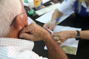 Foto mostra um homem idoso de costas conversando com uma pessoa em uma mesa, onde está sendo atendido.