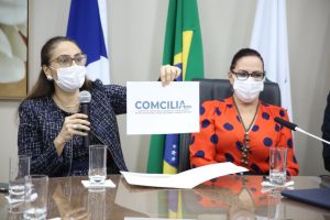 Foto mostra duas mulheres sentadas em uma mesa durante o evento, com uma segurando uma folha com a logomarca do Comcilia enquanto usa a outra mão para segurar um microfone.