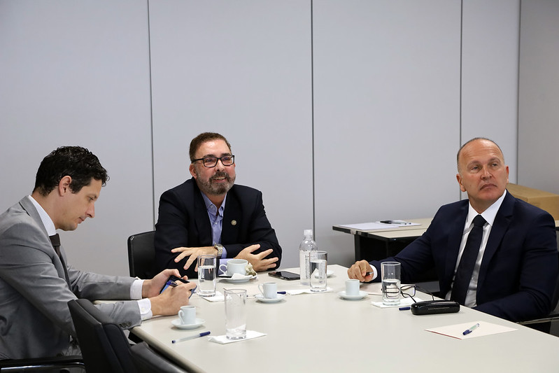 Foto mostra o conselheiro e dois homens sentados em uma mesa, olhando para frente de forma atenta.