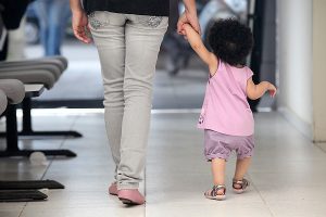Foto mostra uma mulher, da cintura para baixo, e uma criança, ambas de costas e andando de mãos dadas em um corredor.