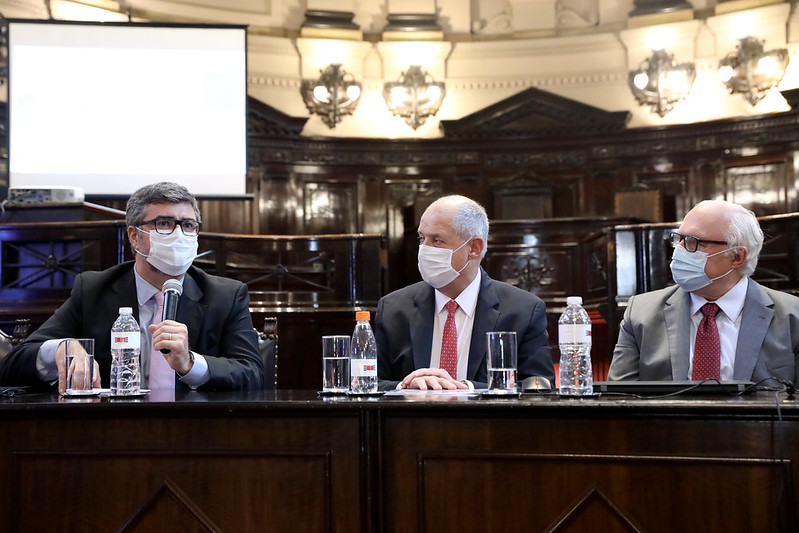 Foto mostra o secretário-geral do CNJ falando na bancada principal do Tribunal do Juri do TJSP. A seu lado, outros dois homens olham atentamente para ele. Ao fundo, é possível ver um telão.