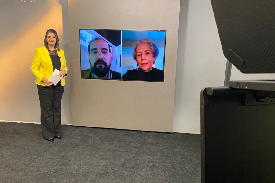 Foto do estúdio do programa com o apresentador posando ao lado do telão onde se vê as pessoas entrevistadas por videoconferência.