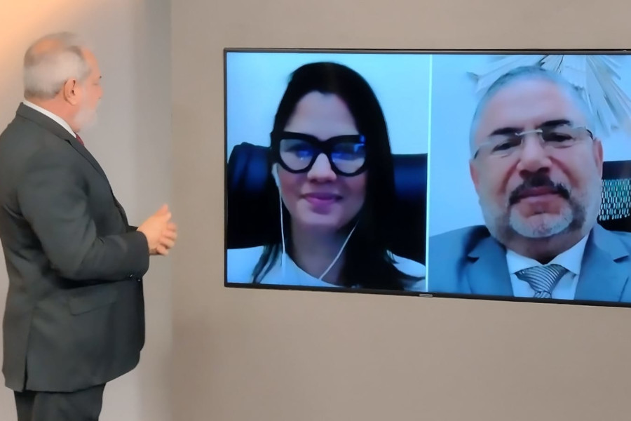 Foto do estúdio do programa com o apresentador posando ao lado do telão onde se vê as pessoas entrevistadas por videoconferência.