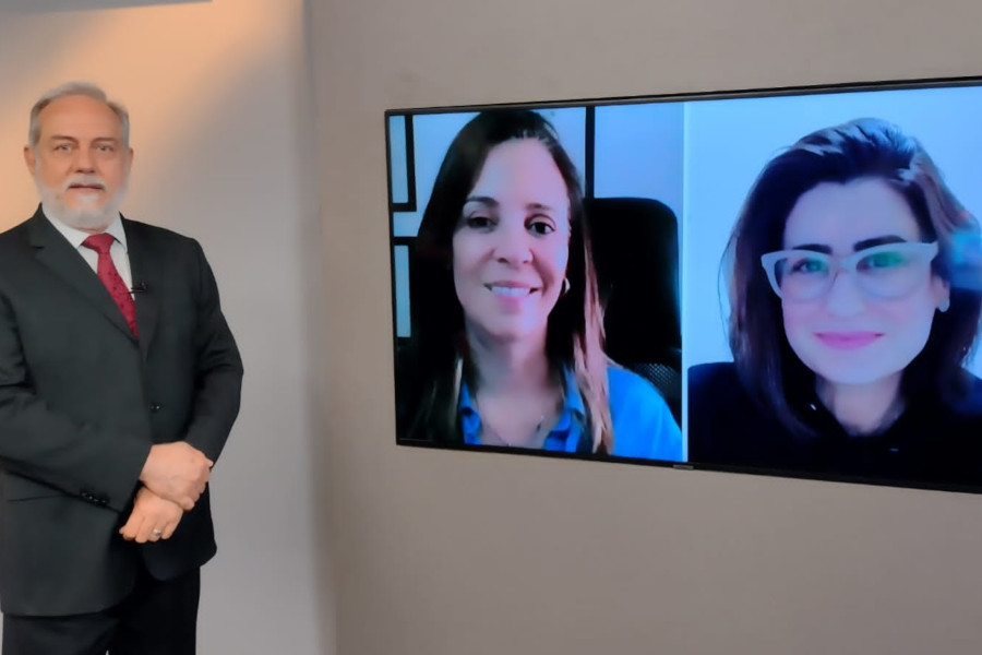 Foto do estúdio do programa com o apresentador posando ao lado do telão onde se vê as pessoas entrevistadas por videoconferência.