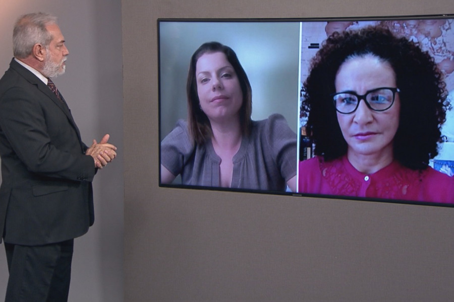 Foto do estúdio do programa com o apresentador posando ao lado do telão onde se vê as pessoas entrevistadas por videoconferência.