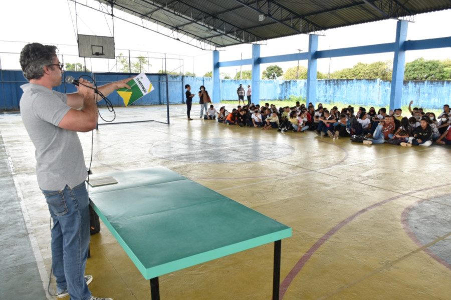 Foto de uma quadra poliesportiva. Em um lado, está de pé o conselheiro, com uma mesa à frente, segurando a Constituição e falando em um microfone. Do outro lado, estão os estudantes participantes do evento.