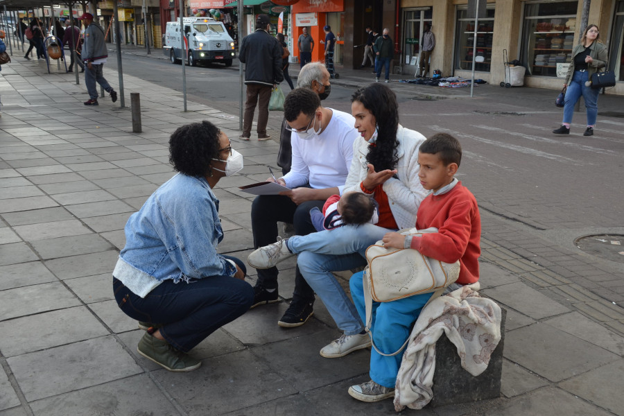 Foto mostra, em um banco de praça em Porto Alegre, um juiz e uma juíza usando máscaras de proteção contra a Covid-19 e ouvindo a dona de casa Ana Paula, que tem um filho sentado a seu lado e um bebê no colo.