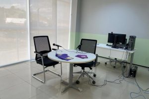 Foto mostra uma sala com uma mesa de escritório com um computador. À frente, tem uma mesa redonda, com duas cadeiras e microfones onde ficam o entrevistador e a criança ou adolescente.