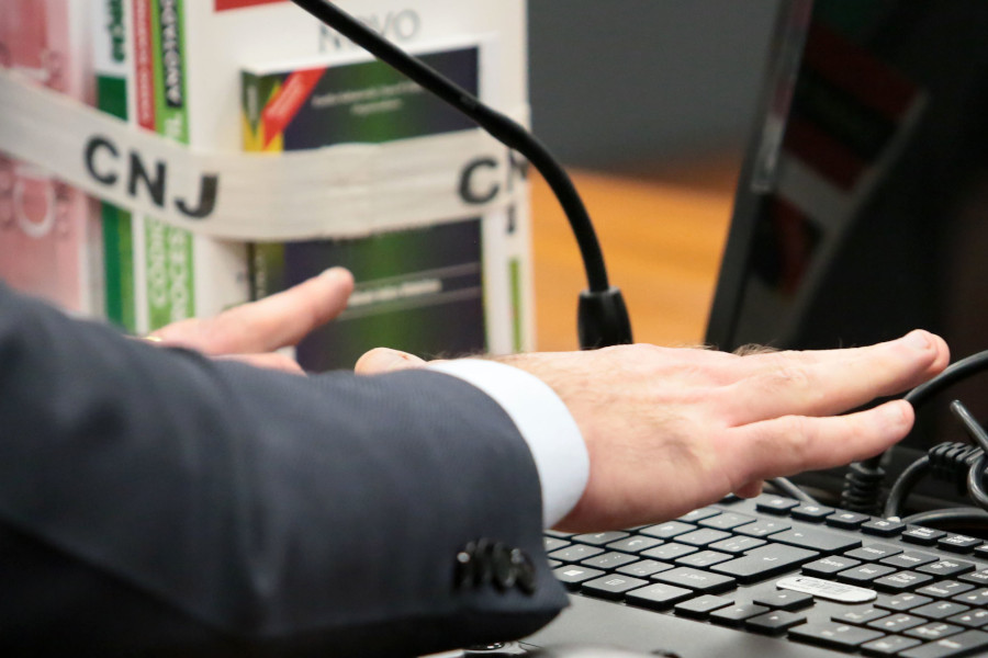Foto mostra a mão de um homem usando um teclado de computador. Ao fundo há alguns livros presos com uma faixa com a marca CNJ.