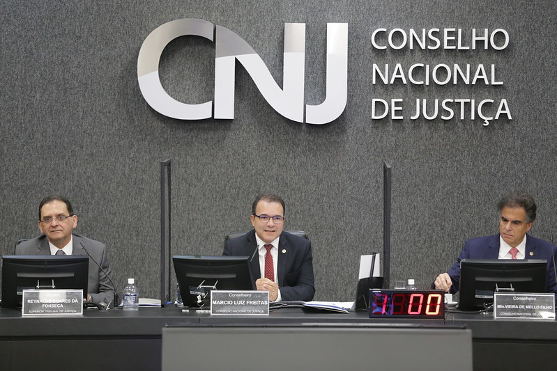 Foto mostra os conselheiros Marcio Freitas e Vieira de Mello e o ministro do STJ Reynaldo Fonseca sentados na bancada principal do Plenário do CNJ durante a abertura do evento.