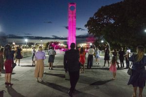 Foto noturna mostra diversas pessoas em uma praça olhando para o Monumento Marco Zero do Equador que está iluminado na cor lilás.