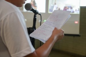 Foto mostra um jovem declamando a poesia que está manuscrita em uma folha de papel.