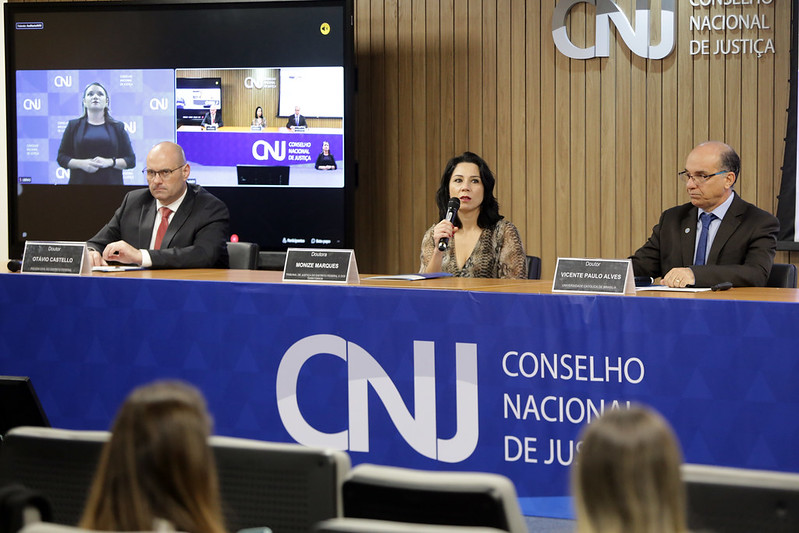 Foto mostra uma mulher e dois homens sentados na bancada principal no palco do Auditório do CNJ durante o encontro.