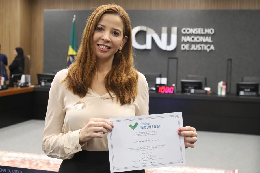 Foto mostra a juíza Déa Marisa Yule em pé, no Plenário do CNJ, segurando o certificado recebido pela premiação.