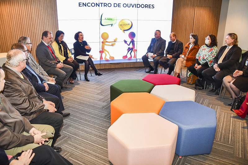 Foto mostra momento da reunião, com as pessoas sentadas em círculo. Ao fundo, em um telão, tem a logomarca do Encontro de Ouvidores.