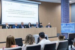 Foto mostra visão da bancada principal do evento, onde se vê, atrás das pessoas, um telão com o texto "Posse dos membros do Conselho Regional da Comunidade da 1ª Vara Regional da Execução Penal e do Conselho da Comunidade do Recife".