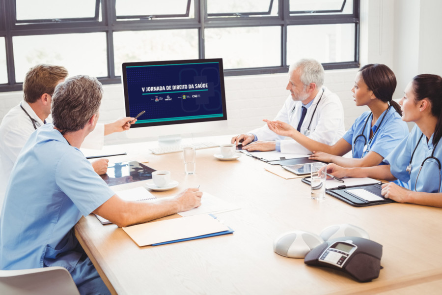 Foto mostra médicos e médicas sentadas em volta de uma mesa e olhando para um monitor, na ponta da mesa, e conversando. No monitor, tem o banner de divulgação do evento.