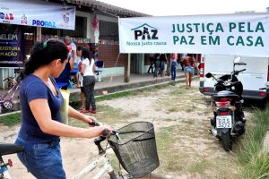 Foto mostra uma mulher de pé, segurando uma bicicleta, olhando para uma escola com mulheres à frente e uma faixa com o texto "Justiça pela Paz em Casa".