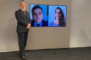 Foto do estúdio do programa com o apresentador posando ao lado do telão onde se vê as pessoas entrevistadas por videoconferência.