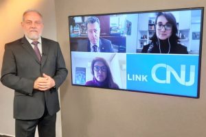 Foto do estúdio do programa com o apresentador posando ao lado do telão onde se vê as pessoas entrevistadas por videoconferência.