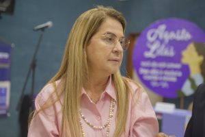 Foto mostra a desembargadora Graça Figueiredo. Ao fundo, uma parede em tom azul escuro e a imagem desfocada de um cartaz em formato redondo, com informações da campanha Agosto Lilás, que também faz alusão ao combate à violência de gênero.