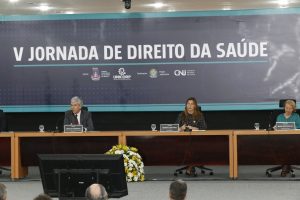 Foto mostra, sentados no palco do evento, o membro do Comitê Executivo do Fonajus, Arnaldo Hossepian, a representante do Hospital Albert Einstein Rogéria Leoni Cruz e a chefe de gabinete da Secretaria de Atenção Especializada do Ministério da Saúde, Maria Inês Gadelha,