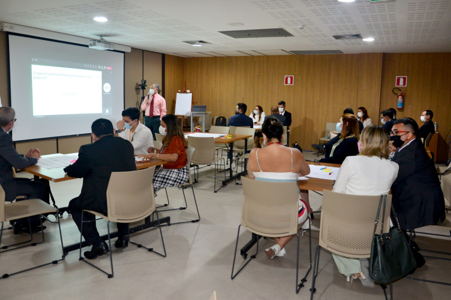 Foto mostra uma sala, com uma pessoa à frente, em pé, fazendo uma apresentação em um telão e as demais pessoas sentadas em grupos em mesas de quatro lugares.