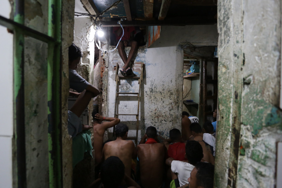 Foto mostra uma cela com presos amontoados - sentados no chão e de costas -, fiações elétricas expostas, paredes corroídas e outros problemas.