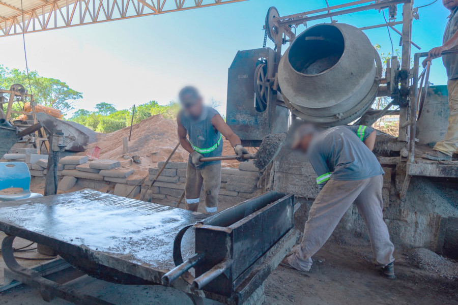 Foto mostra dois homens em frente à betoneira, colocando concreto em um molde retangular de uma tampa. Ao fundo estão insumos, como cimento, areia e brita. Na imagem, o dia está com o céu bem azulado e o sol está forte.