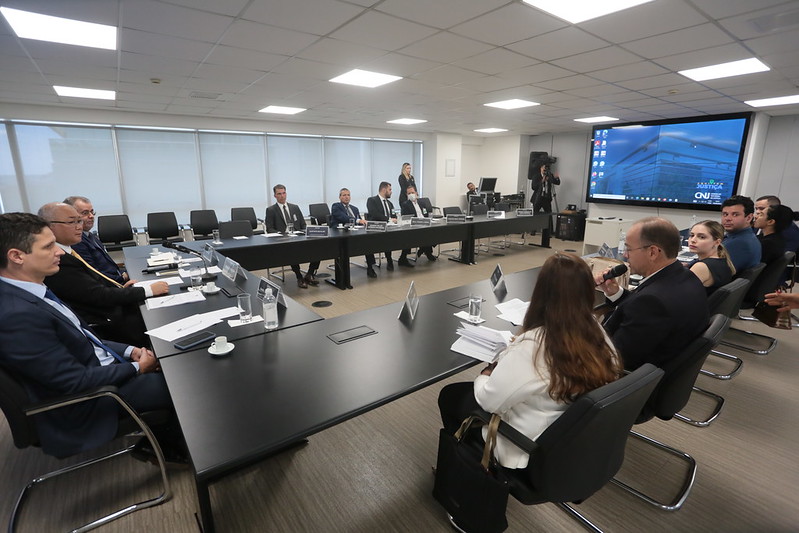 Foto mostra momento da reunião, com as pessoas sentadas em volta de uma grande mesa em U, com um telão ao fundo.