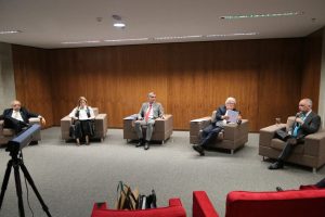 Foto mostra momento do evento no auditório da Enfam, com participantes da mesa de abertura sentados em poltronas no palco.