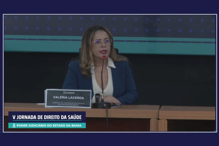 Foto mostra a juíza sentada na mesa do palco do evento e fazendo sua apresentação.
