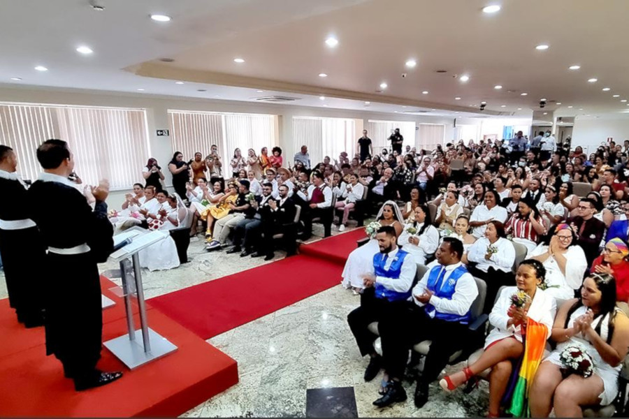 Foto mostra momento da cerimônia, com o auditório cheio, os casais nas primeiras cadeiras e os dois magistrados em pé falando ao microfone.