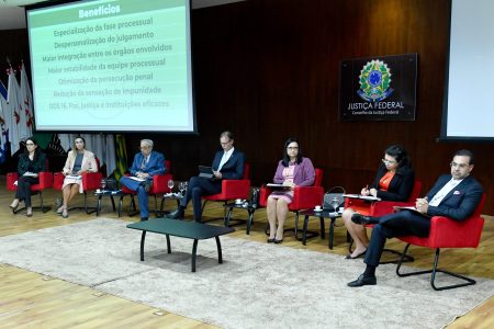 Foto mostra palco, com um telão ao fundo, e os participantes sentados em cadeiras.