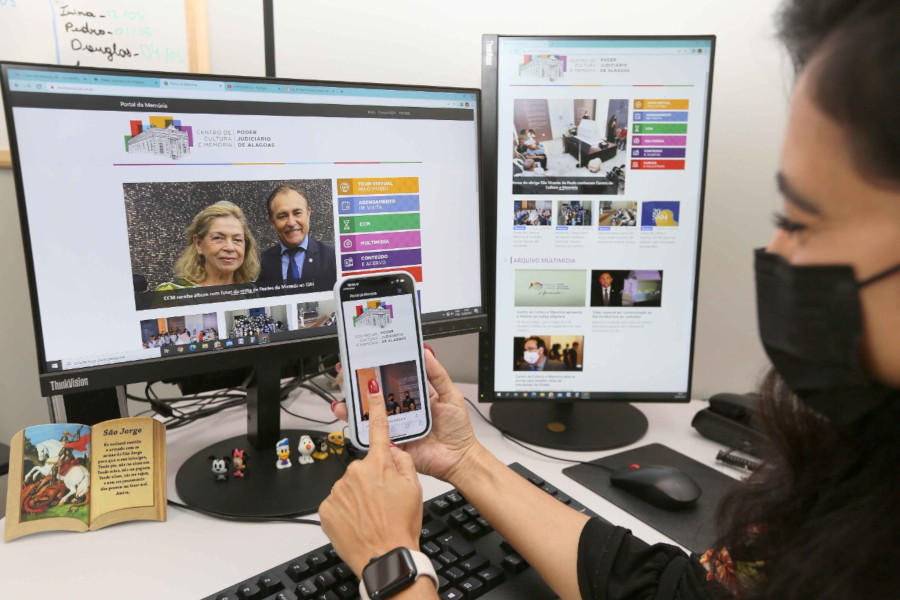 Foto mostra uma mulher com o celular na mão, apontando para a tela. Atrás, na mesa de trabalho dela, tem dois monitores. Celular e monitores estão com a tela de acesso do Portal da Memória.