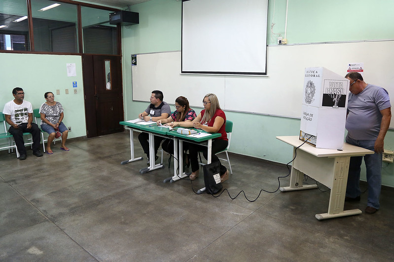 Foto mostra uma seção eleitoral em uma sala de aula, onde se vê um homem em pé votando na cabine, os mesários sentados atrás de mesas e pessoas aguardando para votar também sentadas.