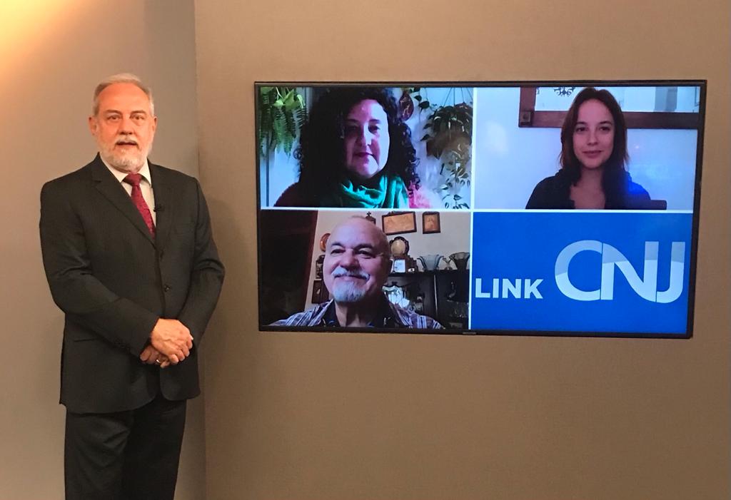 Foto do estúdio do programa com o apresentador posando ao lado do telão onde se vê as pessoas entrevistadas por videoconferência.