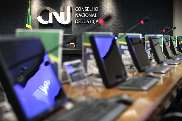 Imagem da mesa plenária do CNJ, em primeiro plano mesa composta por computadores, microfones e livros, ao fundo painel de tom cinza escuro com a logo do Conselho Nacional de Justiça e bandeira do Brasil.