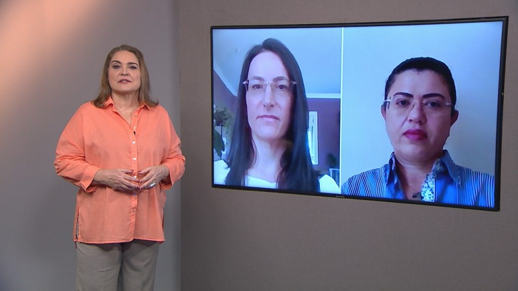 Foto do estúdio do programa com o apresentador posando ao lado do telão onde se vê as pessoas entrevistadas por videoconferência.