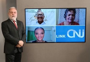 Foto do estúdio do programa com o apresentador posando ao lado do telão onde se vê as pessoas entrevistadas por videoconferência.