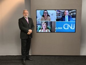 Foto do estúdio do programa com o apresentador posando ao lado do telão onde se vê as pessoas entrevistadas por videoconferência.