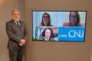 Foto do estúdio do programa com o apresentador posando ao lado do telão onde se vê as pessoas entrevistadas por videoconferência.