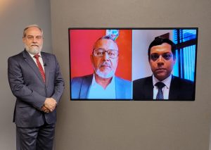 Foto do estúdio do programa com o apresentador posando ao lado do telão onde se vê as pessoas entrevistadas por videoconferência.