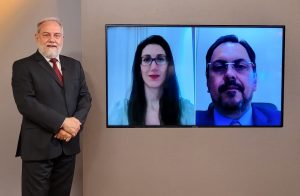 Foto do estúdio do programa com o apresentador posando ao lado do telão onde se vê as pessoas entrevistadas por videoconferência.