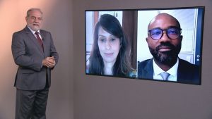 Foto do estúdio do programa com o apresentador posando ao lado do telão onde se vê as pessoas entrevistadas por videoconferência.