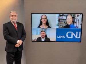 Foto do estúdio do programa com o apresentador posando ao lado do telão onde se vê as pessoas entrevistadas por videoconferência.