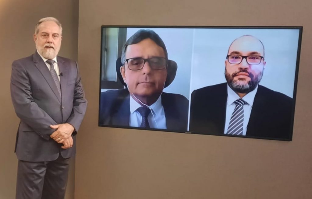 Foto do estúdio do programa com o apresentador posando ao lado do telão onde se vê as pessoas entrevistadas por videoconferência.