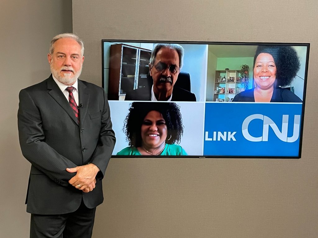 Foto do estúdio do programa com o apresentador posando ao lado do telão onde se vê as pessoas entrevistadas por videoconferência.