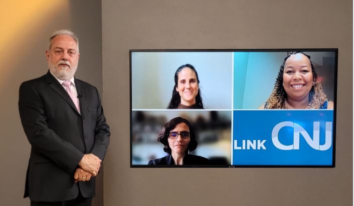 Foto do estúdio do programa com o apresentador posando ao lado do telão onde se vê as pessoas entrevistadas por videoconferência.